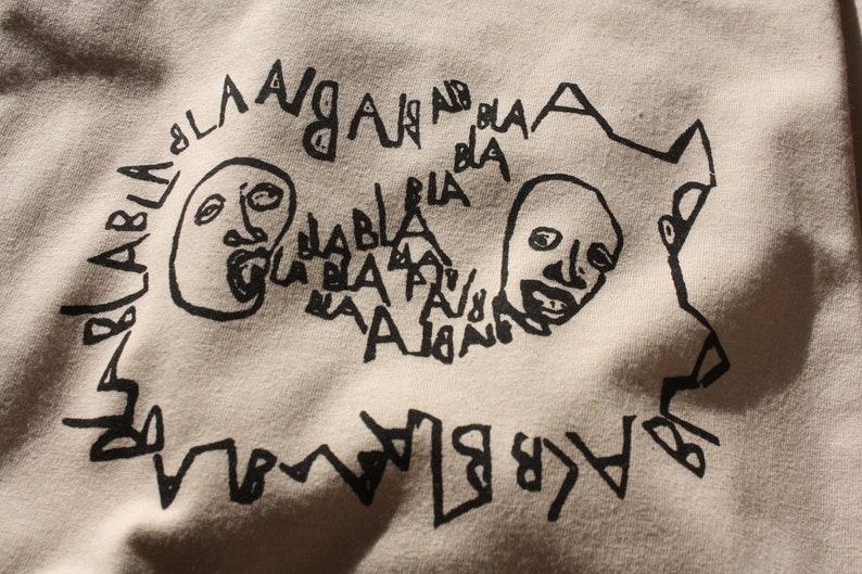 May include: Black and white graphic print on a beige fabric featuring two faces with open mouths and the words "bla bla bla" and "angan" repeated around the faces.
