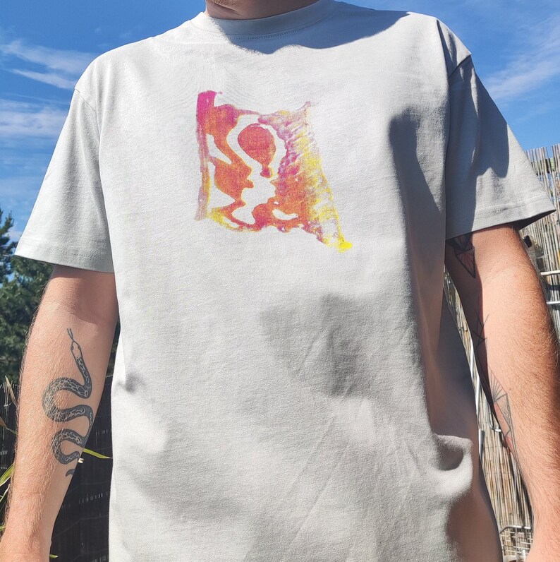 May include: A light gray t-shirt featuring a colorful abstract design in shades of pink, yellow, and orange. The graphic is centered on the chest. The shirt has short sleeves and a crew neck.