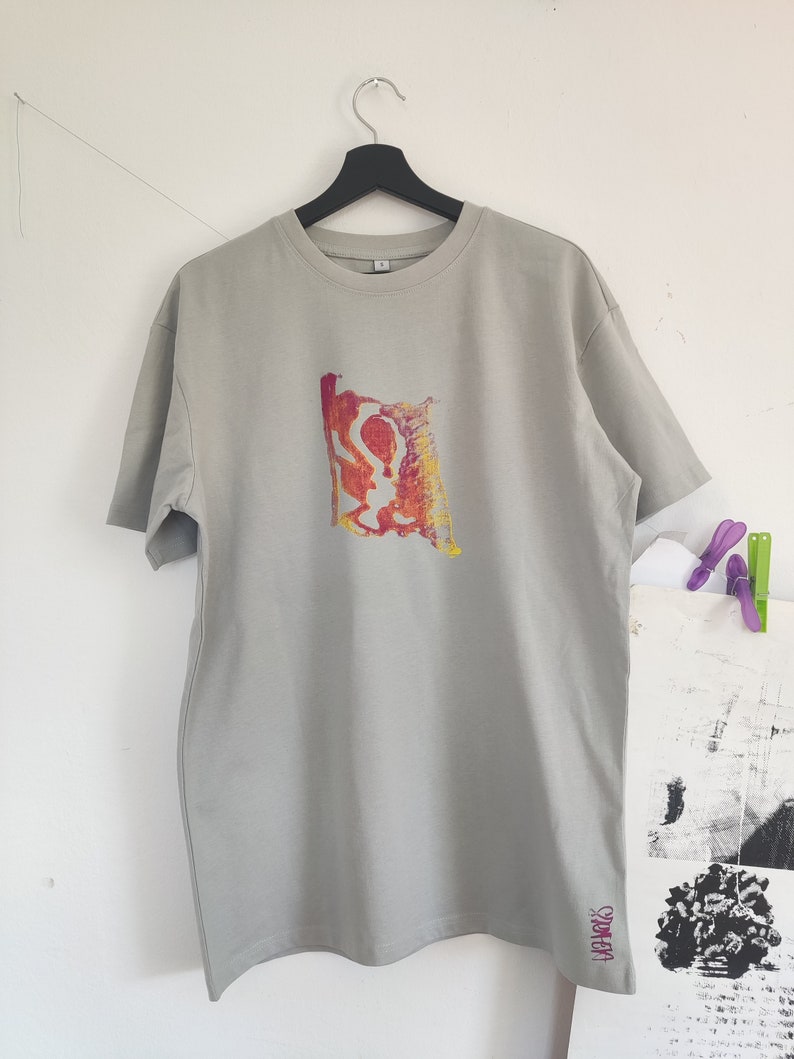 May include: A light gray t-shirt with a colorful abstract design on the front. The design features red, yellow, and orange hues. The word "SPARKS" is printed in pink on the lower right side of the shirt.