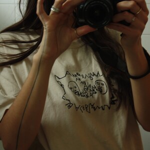 May include: A person wearing a white t-shirt with a black graphic design of two faces and the text "ALGEBRA HABLA ALGEBRA HABLA". The person is holding a camera up to a mirror.
