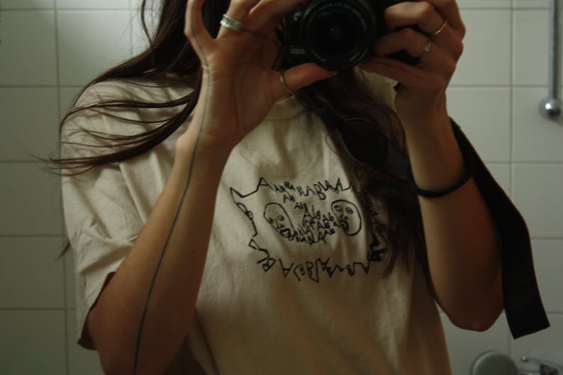 May include: A person wearing a white t-shirt with a black graphic design of two faces and the text "ALGEBRA HABLA ALGEBRA HABLA". The person is holding a camera up to a mirror.