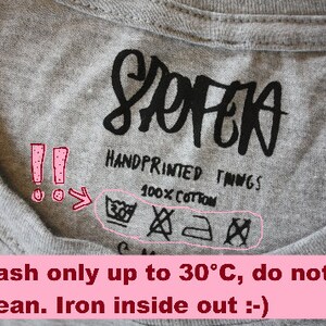 May include: Close-up of a gray t-shirt label with the word "SOPEA" in black script. The label reads "HANDPRINTED THINGS" and "100% COTTON." Washing instructions include a symbol for washing at 30&deg;C, do not dry clean, and iron inside out.