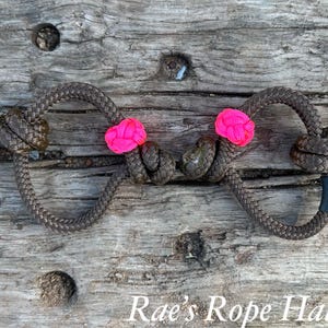 May include: A brown rope halter featuring two bright neon pink knots positioned centrally and four additional brown knots at the ends. A small black tag with white text reading Haltors is attached to the right side of the braided rope structure.