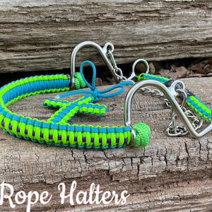May include: A green and blue braided rope halter with a silver bit. The halter is on a wooden surface. Raes Rope Halters.