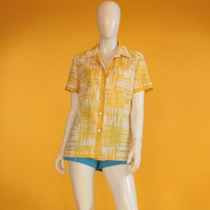 Op de afbeelding: Een geel en wit gepatroneerd shirt met korte mouwen en een knoopsluiting. Het shirt is op een wit mannequins met een blauwe broek.