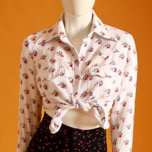 May include: A white button-down shirt with a floral pattern of red, blue, and green flowers, tied at the waist. Paired with black pants covered in red cherry designs. The outfit is displayed on a mannequin.
