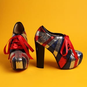 Puede incluir: Par de zapatos de tacón alto con plataforma con un diseño de lentejuelas de la Union Jack en rojo, blanco, azul y dorado. Los zapatos tienen tacones negros y cordones de satén rojos, sobre un fondo amarillo.