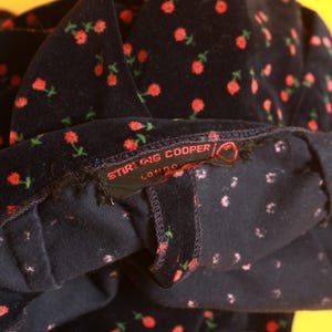May include: Close-up of a dark navy fabric with a repeating pattern of small red roses and green stems. The label reads "STIRLING COOPER LONDON". The fabric appears to be a soft velvet or corduroy.