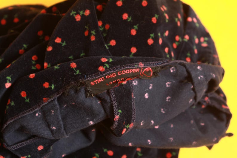 May include: Close-up of a dark navy fabric with a repeating pattern of small red roses and green stems. The label reads "STIRLING COOPER LONDON". The fabric appears to be a soft velvet or corduroy.