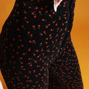 May include: Black velvet pants with a repeating pattern of small red roses and green leaves. The high-waisted pants are paired with a white top with a floral pattern. The pants are a stylish and unique piece of clothing.