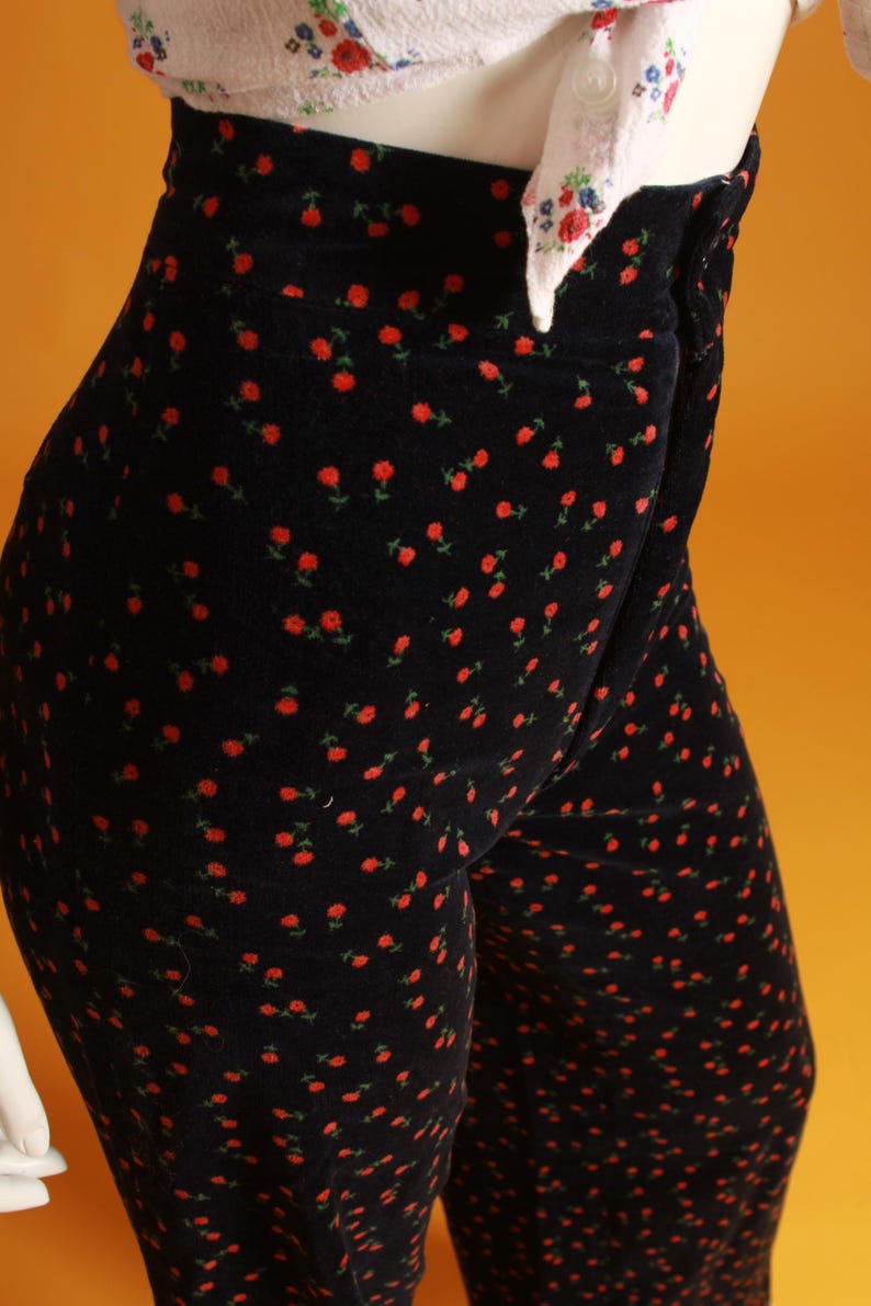 May include: Black velvet pants with a repeating pattern of small red roses and green leaves. The high-waisted pants are paired with a white top with a floral pattern. The pants are a stylish and unique piece of clothing.