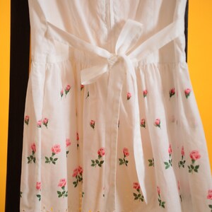 Puede incluir: Vestido blanco con estampado de rosas. El vestido tiene una cintura con lazo en la espalda y un dobladillo festoneado. Rosas rosas con hojas verdes est&aacute;n impresas en la tela. El vestido cuelga sobre una superficie negra sobre un fondo amarillo.
