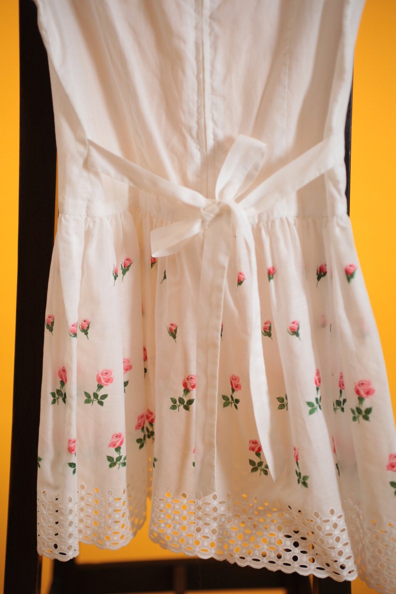 Puede incluir: Vestido blanco con estampado de rosas. El vestido tiene una cintura con lazo en la espalda y un dobladillo festoneado. Rosas rosas con hojas verdes est&aacute;n impresas en la tela. El vestido cuelga sobre una superficie negra sobre un fondo amarillo.