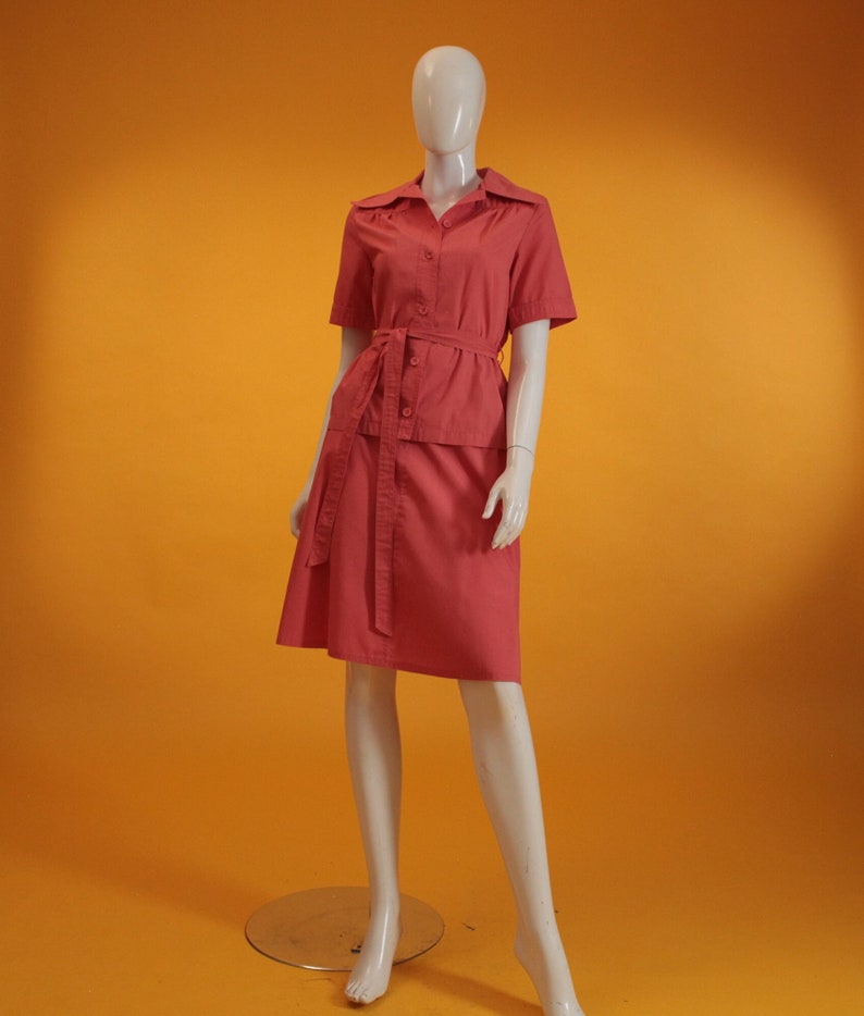 May include: A coral pink short-sleeved collared blouse with a matching skirt. The blouse has a belt and buttons down the front. The skirt is a-line and falls to the knees.