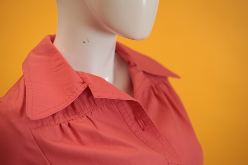 May include: Close-up of a coral pink button-down shirt with a pointed collar. The shirt is made of a soft, smooth fabric.