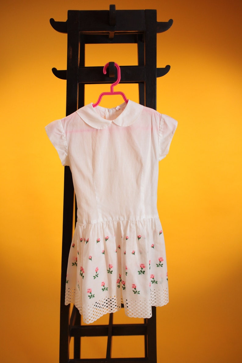 Puede incluir: Un vestido blanco con cuello Peter Pan y mangas cortas cuelga de una percha rosa. El vestido presenta un estampado floral de rosas rosas en la falda y un dobladillo festoneado. El vestido se exhibe en un perchero de madera negro sobre un fondo amarillo.