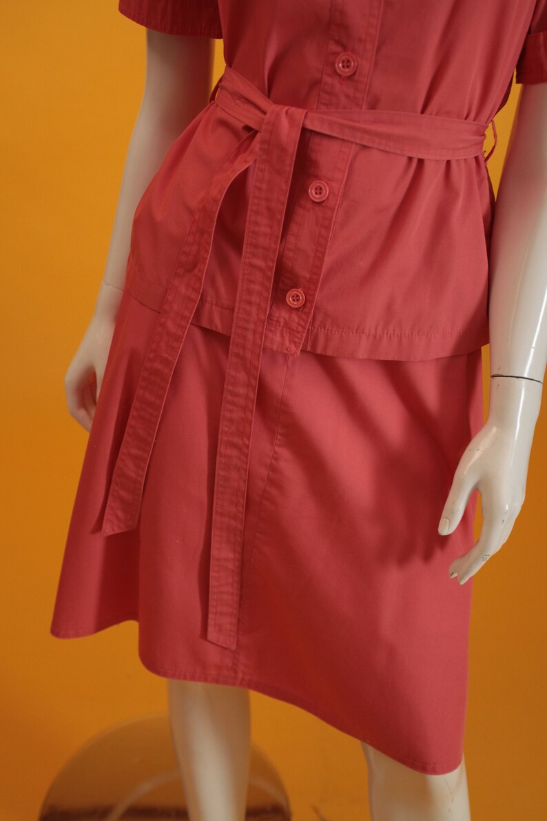 May include: A coral pink short-sleeved dress with a matching belt. The dress has a button-up front and a skirt with a slight A-line shape.