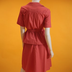 May include: A coral pink dress with a short-sleeved top and a skirt. The top has a collar and a belt at the waist.