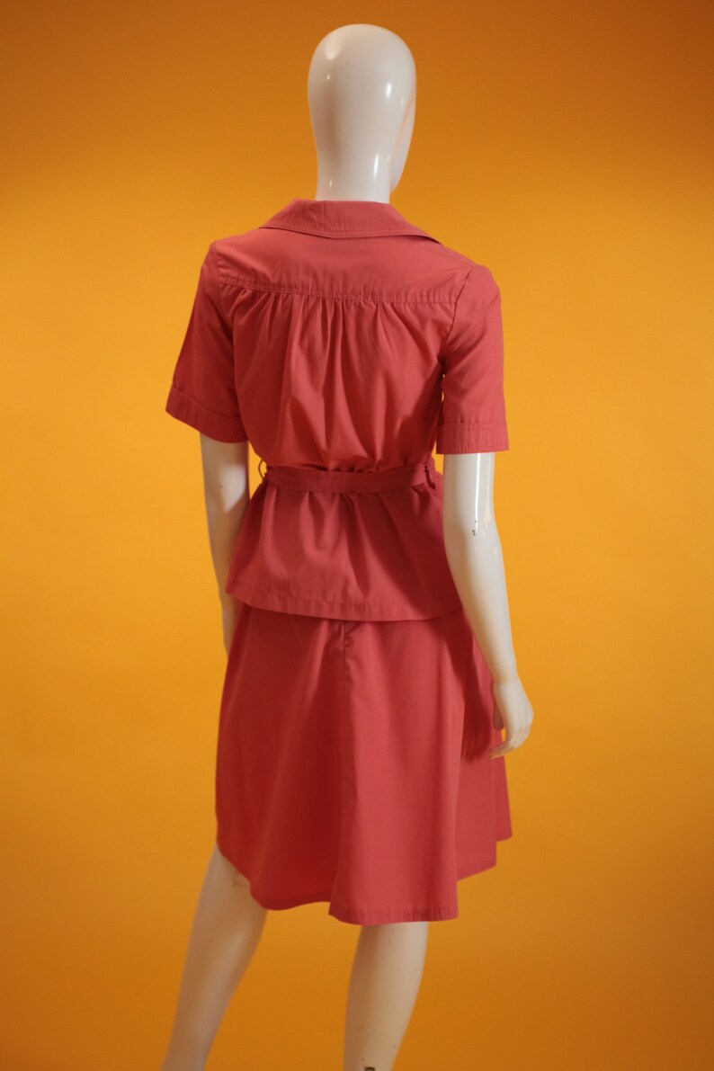 May include: A coral pink dress with a short-sleeved top and a skirt. The top has a collar and a belt at the waist.