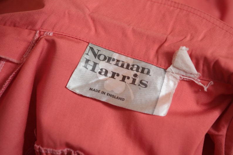 May include: A white label with black text that reads "Norman Harris" and "Made in England". The label is attached to a coral colored fabric.