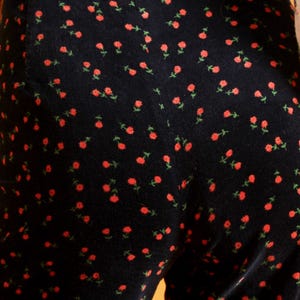 May include: Close-up of a black jumpsuit with a repeating pattern of small red roses and green stems. The jumpsuit is paired with a light-colored blouse with a floral print. The overall style is vintage-inspired.