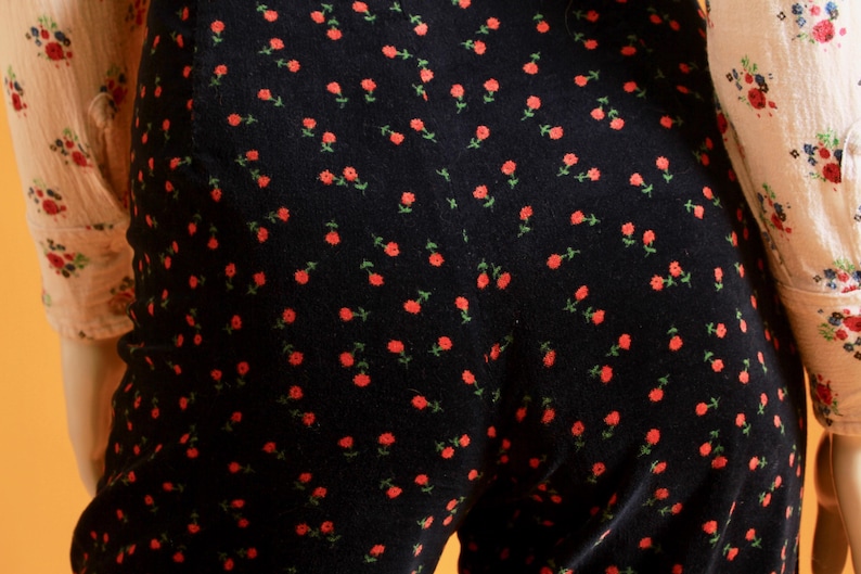 May include: Close-up of a black jumpsuit with a repeating pattern of small red roses and green stems. The jumpsuit is paired with a light-colored blouse with a floral print. The overall style is vintage-inspired.