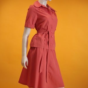 May include: A coral pink short-sleeved collared blouse with a matching skirt. The blouse has a tie belt and button closure. The skirt is a-line and has a tie belt.
