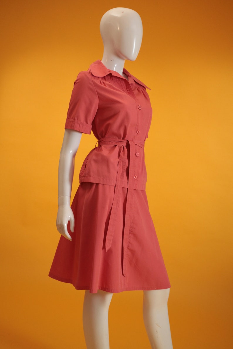 May include: A coral pink short-sleeved collared blouse with a matching skirt. The blouse has a tie belt and button closure. The skirt is a-line and has a tie belt.