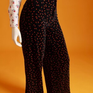 May include: Black velvet pants with a red floral pattern. The high-waisted pants are paired with a white blouse with a floral print. The pants have a wide leg design, and the outfit is displayed on a mannequin.