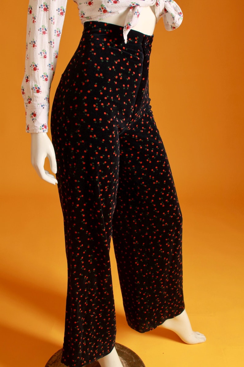 May include: Black velvet pants with a red floral pattern. The high-waisted pants are paired with a white blouse with a floral print. The pants have a wide leg design, and the outfit is displayed on a mannequin.