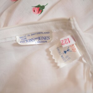 Puede incluir: Primer plano de una prenda blanca con un delicado estampado floral de rosas rosas y hojas verdes. La etiqueta dice "MADE IN SWITZERLAND FOR DICKINS AND JONES LONDON". Se ven etiquetas adicionales.