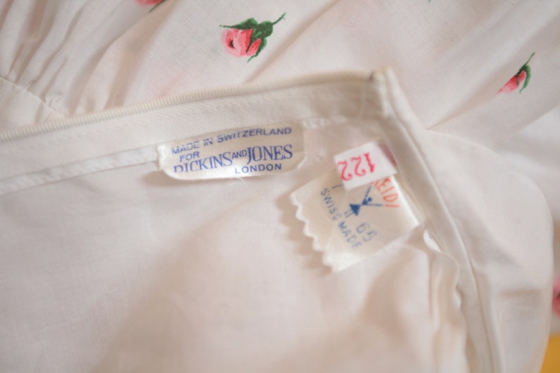 Puede incluir: Primer plano de una prenda blanca con un delicado estampado floral de rosas rosas y hojas verdes. La etiqueta dice "MADE IN SWITZERLAND FOR DICKINS AND JONES LONDON". Se ven etiquetas adicionales.