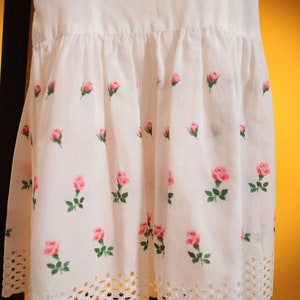 Puede incluir: Vestido blanco con estampado de rosas. El vestido presenta una falda fruncida con rosas rosas y hojas verdes. El dobladillo tiene un borde decorativo de estilo ojal. El vestido est&aacute; colgado en una silla negra.