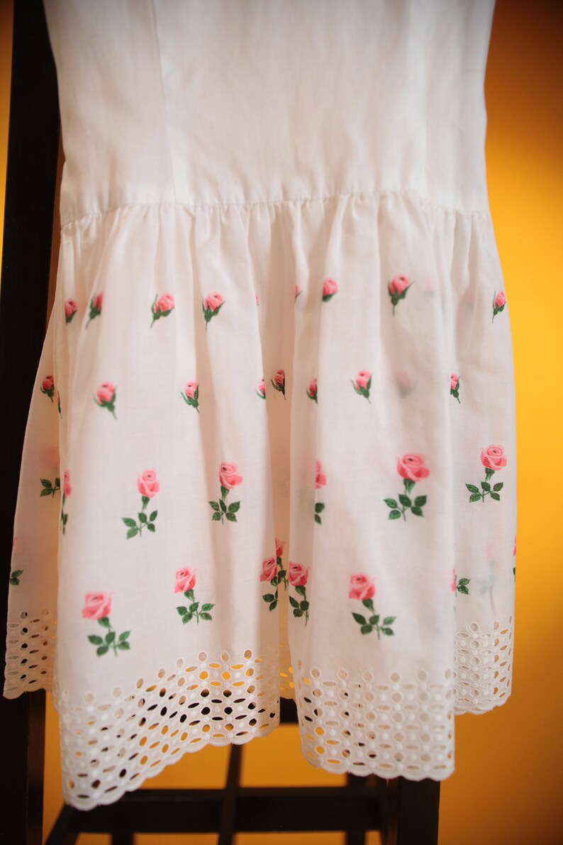 Puede incluir: Vestido blanco con estampado de rosas. El vestido presenta una falda fruncida con rosas rosas y hojas verdes. El dobladillo tiene un borde decorativo de estilo ojal. El vestido est&aacute; colgado en una silla negra.