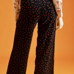 May include: Black wide-leg pants with a repeating pattern of small red flowers. The pants are paired with a cream-colored blouse with a floral print. The outfit is displayed on a mannequin against an orange background.