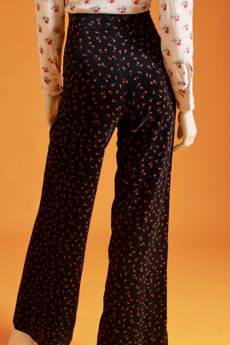 May include: Black wide-leg pants with a repeating pattern of small red flowers. The pants are paired with a cream-colored blouse with a floral print. The outfit is displayed on a mannequin against an orange background.