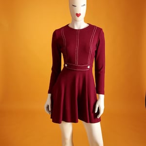 Könnte beinhalten: Burgunderfarbenes Kleid mit langen Ärmeln und ausgestelltem Rock. Das Kleid hat weiße Steppdetails am Oberteil und an der Taille, mit zwei weißen Knöpfen. Das Kleid wird auf einer Schaufensterpuppe vor orangefarbenem Hintergrund präsentiert.