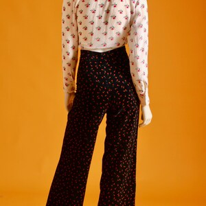 May include: A mannequin is wearing a vintage outfit. The ensemble includes a white button-down shirt with a small red floral pattern and high-waisted, wide-leg black pants with a red dot pattern. The outfit is set against an orange background.