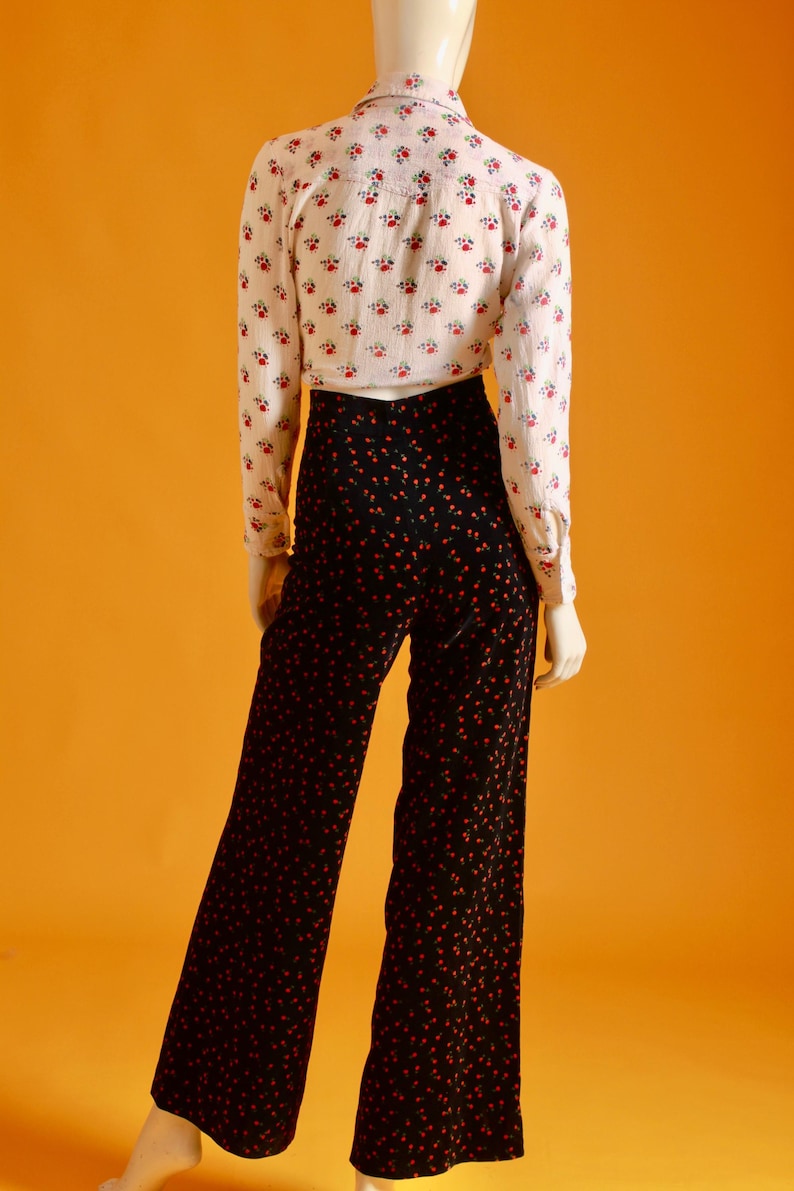May include: A mannequin is wearing a vintage outfit. The ensemble includes a white button-down shirt with a small red floral pattern and high-waisted, wide-leg black pants with a red dot pattern. The outfit is set against an orange background.