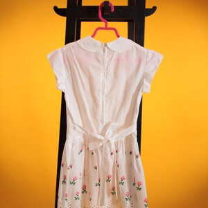 Puede incluir: Vestido blanco con cuello Peter Pan, mangas cortas y cintura con lazo. El vestido presenta un estampado floral de rosas y un dobladillo festoneado. Cuelga de un estante de madera negro sobre un fondo amarillo.
