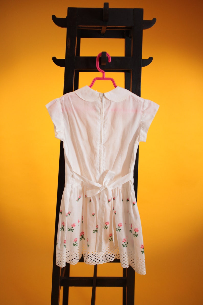 Puede incluir: Vestido blanco con cuello Peter Pan, mangas cortas y cintura con lazo. El vestido presenta un estampado floral de rosas y un dobladillo festoneado. Cuelga de un estante de madera negro sobre un fondo amarillo.