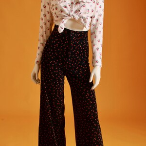 May include: A white button-down shirt with a floral pattern is tied at the waist, paired with black wide-leg pants adorned with a red floral design. The outfit is displayed on a mannequin against an orange background.