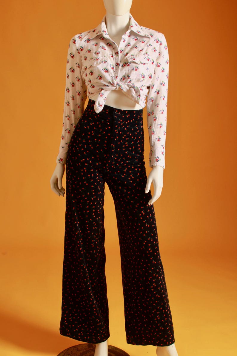 May include: A white button-down shirt with a floral pattern is tied at the waist, paired with black wide-leg pants adorned with a red floral design. The outfit is displayed on a mannequin against an orange background.