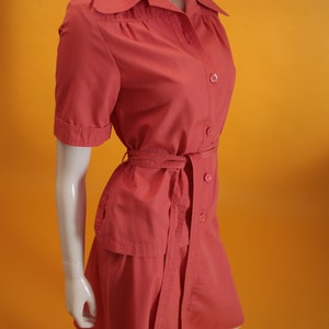 May include: A coral pink short-sleeved dress with a collared neckline, button closure, and a matching belt. The dress has a slightly A-line silhouette and a skirt that falls just above the knee.