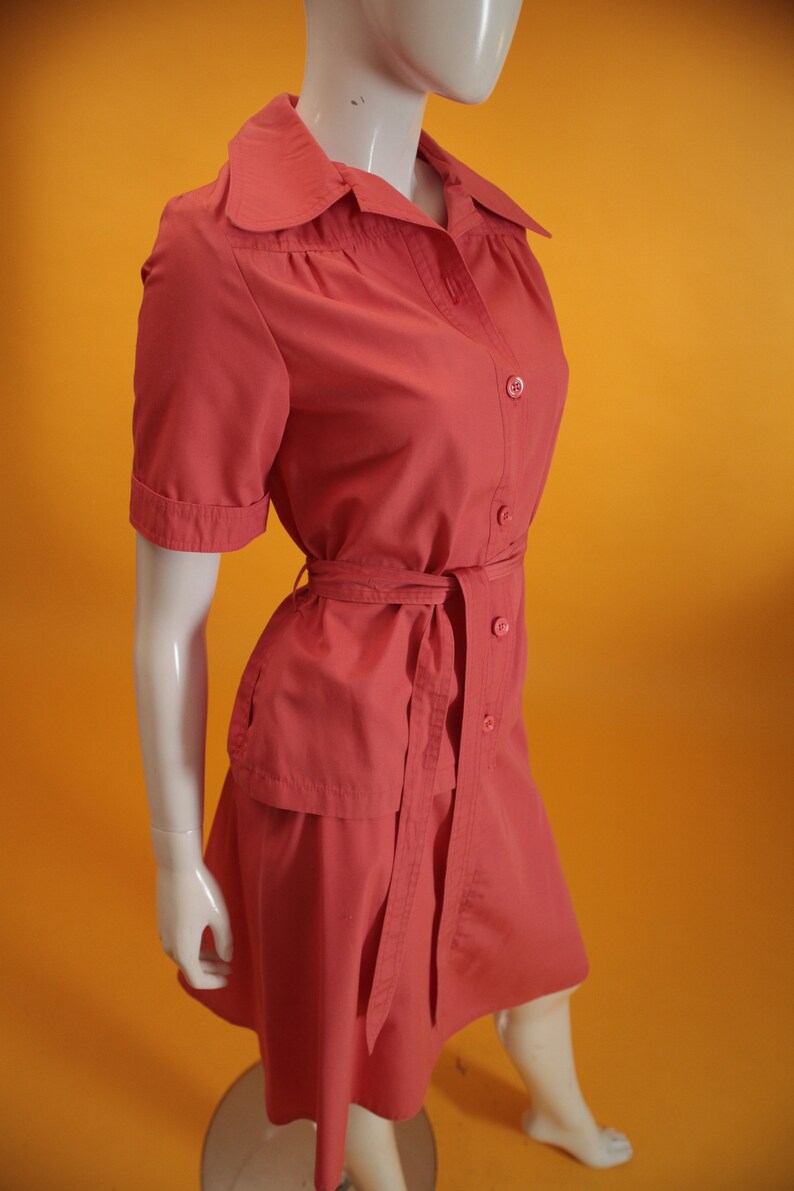 May include: A coral pink short-sleeved dress with a collared neckline, button closure, and a matching belt. The dress has a slightly A-line silhouette and a skirt that falls just above the knee.