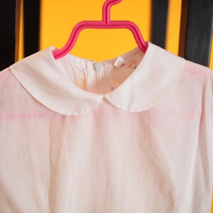 Puede incluir: Un vestido blanco con cuello Peter Pan y mangas cortas cuelga de una percha rosa. El vestido tiene un dise&ntilde;o sencillo y una tela delicada, ligeramente transparente. El cuello es de un blanco n&iacute;tido, contrastando con el color blanquecino del vestido.