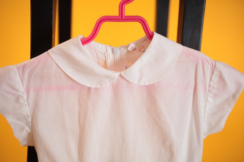Puede incluir: Un vestido blanco con cuello Peter Pan y mangas cortas cuelga de una percha rosa. El vestido tiene un dise&ntilde;o sencillo y una tela delicada, ligeramente transparente. El cuello es de un blanco n&iacute;tido, contrastando con el color blanquecino del vestido.