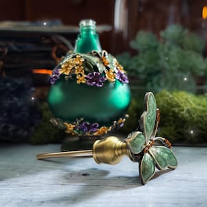 May include: A green glass bottle with a decorative floral design and a butterfly-shaped bottle stopper. The stopper is green with orange accents. The bottle has gold and purple floral accents.