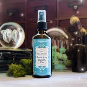 Op de afbeelding: Een bruine glazen spuitfles met een zwarte dop en een lichtblauw etiket. Op het etiket staat "Claire's Lily of the Valley Smudge Spray Dawn Over Craigh na Dun" en "100 ml".