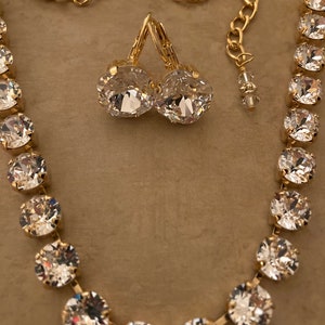 May include: A gold necklace and earrings set with clear crystal stones. The necklace is a simple chain with a row of round crystals. The earrings are dangle earrings with a single round crystal.